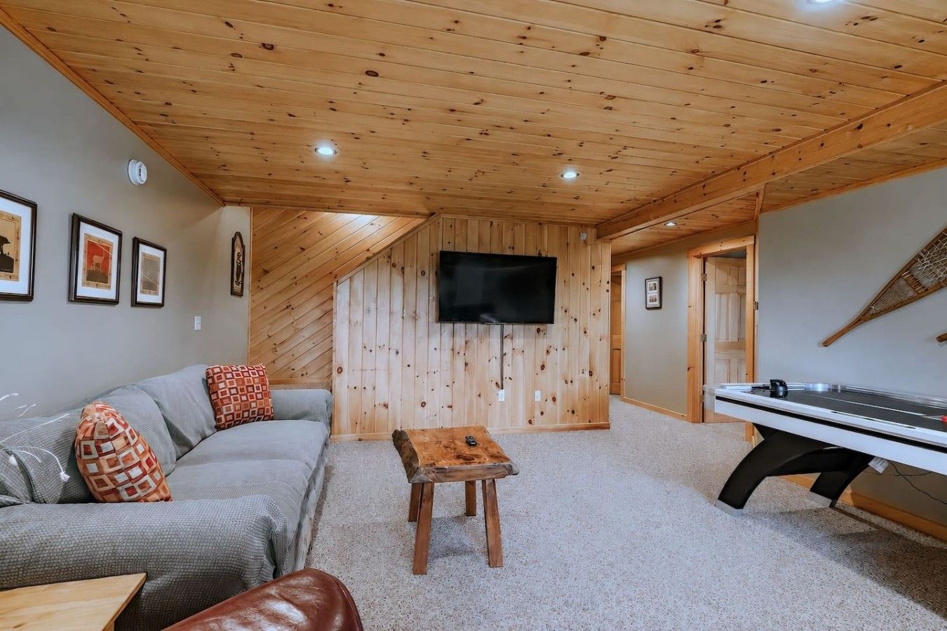 Ein gemütliches Zimmer mit Holzboden, einem grauen Sofa, einem kleinen Holztisch, einem Billardtisch und einem an der Wand befestigten Fernseher.