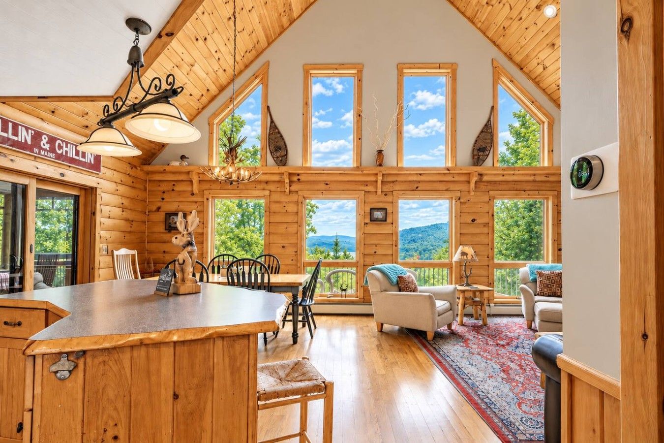 A wooden cabin interior with a dining area, a living room with a sofa, and large windows offering a view of the outdoors.