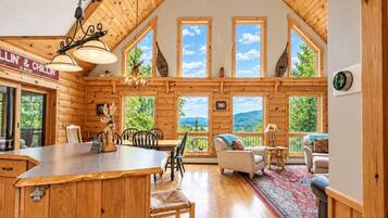 A wooden cabin interior with a dining area, a living room with a sofa, and large windows offering a view of the outdoors.