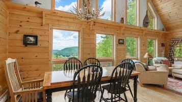 Interior de una cabaña de madera con una mesa de comedor, sillas, un candelabro y ventanas amplias que dan a vistas del exterior.