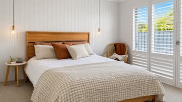 A bedroom with a wooden headboard, a checkered bedspread, and a view of greenery through the window blinds.