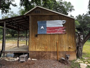 A wooden shed with a flag on the side, a wooden deck, and a small outdoor area with rocks and logs. - PRIVATE COZY RV FOR RENT; ONLY 2 RVS ON PROPERTY, HOT TUB, CHARCOAL GRILL;SERENE (Canyon Lake)