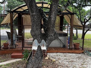 A tree with a metal structure resembling wings attached to its trunk, a covered outdoor seating area, and a fenced patio. - PRIVATE COZY RV FOR RENT; ONLY 2 RVS ON PROPERTY, HOT TUB, CHARCOAL GRILL;SERENE (Canyon Lake)