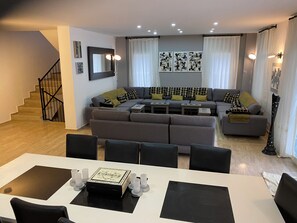 A modern living room with a sectional sofa, a dining table with chairs, and a staircase.