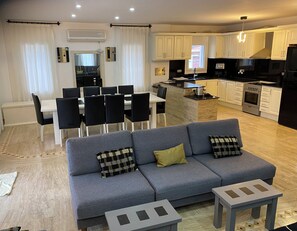 A modern living room with a grey sofa, a dining area with chairs, and a kitchen with white cabinets and appliances.
