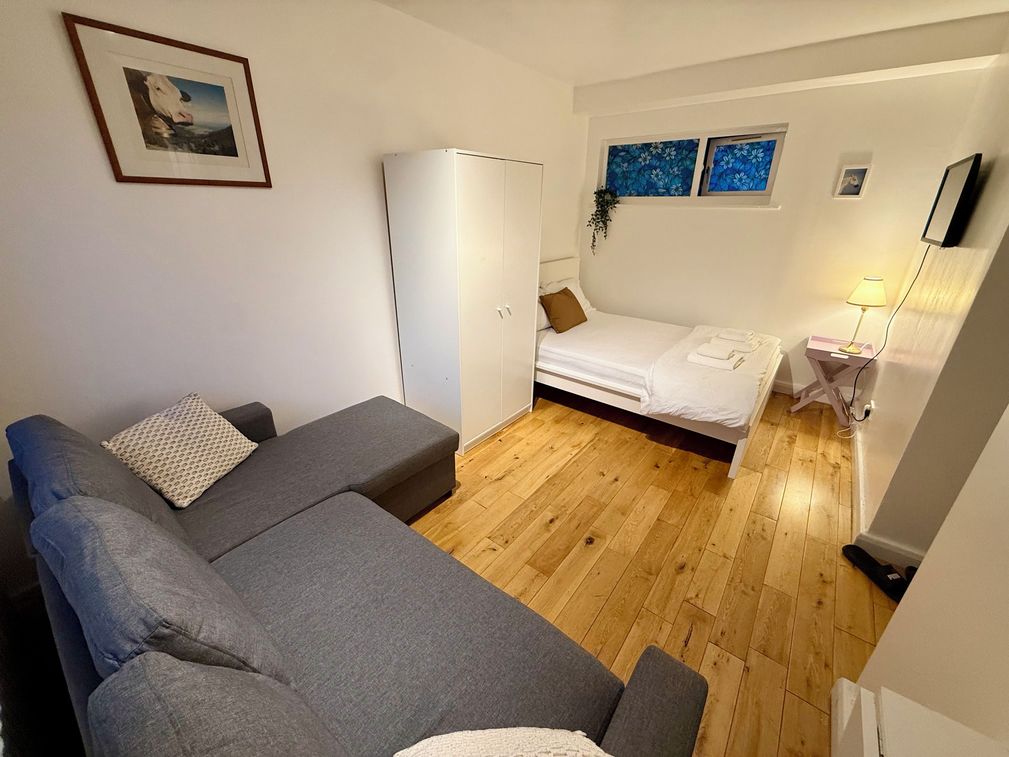 Une pièce de taille modeste, avec un canapé gris, un lit doté d’un matelas blanc, une armoire et une petite table de chevet avec une lampe.
