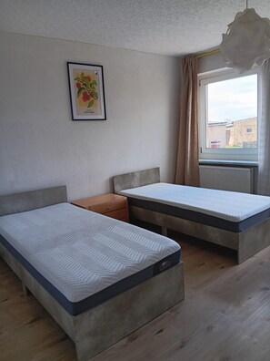 Ein Zimmer mit zwei Betten, einem kleinen Tisch, einem gerahmten Bild an der Wand und einem Fenster mit Vorhängen.