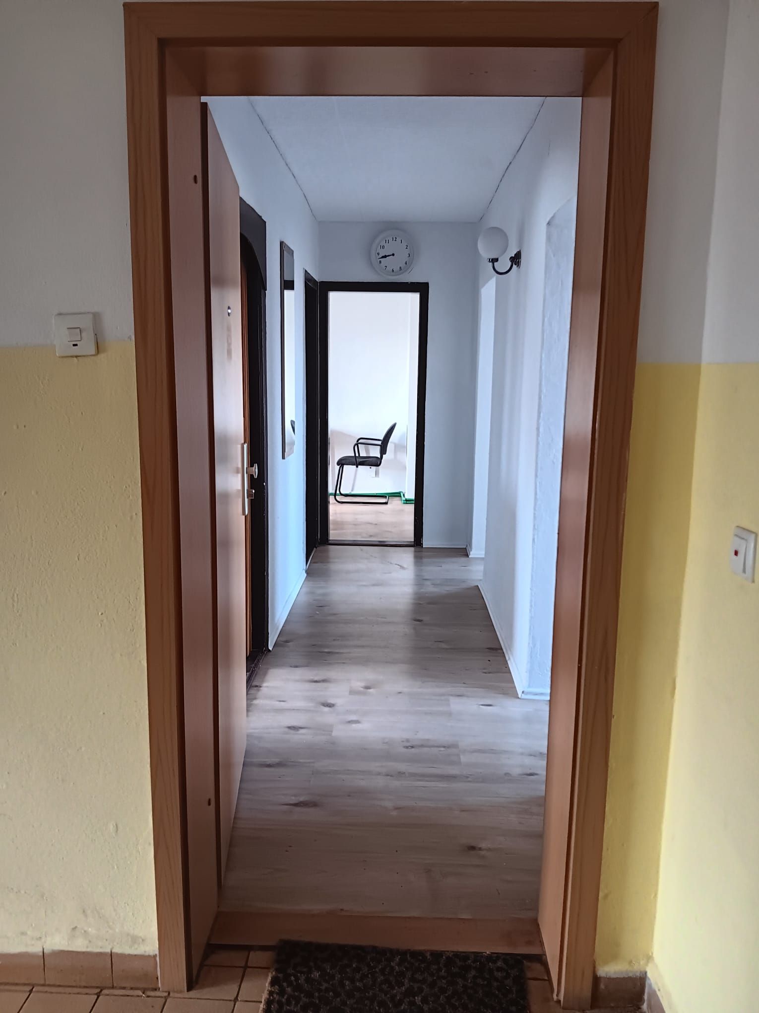 A hallway with a wooden floor, a clock on the wall, and a chair in the distance.