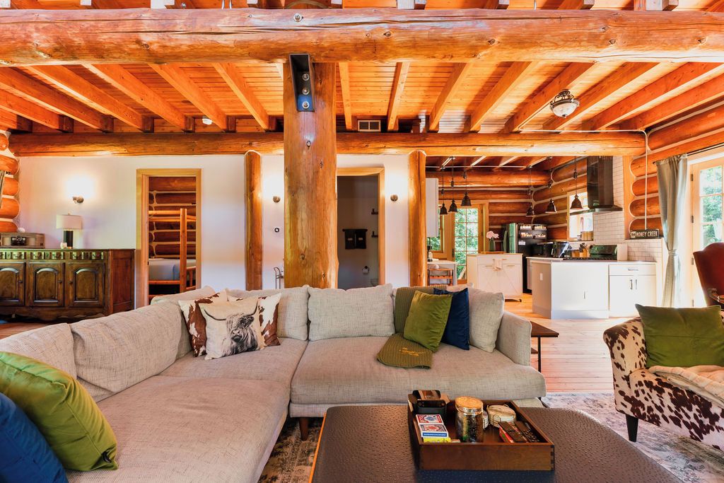 Ein Holzblockhaus-Interieur mit einem großen, L-förmigen Ecksofa, einem Holztisch und einem durch einen offenen Durchgang sichtbaren Küchenbereich.
