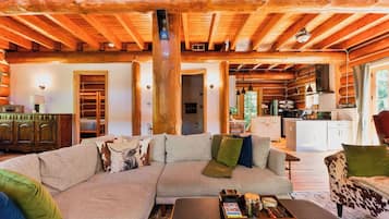 Ein Holzblockhaus-Interieur mit einem großen, L-förmigen Ecksofa, einem Holztisch und einem durch einen offenen Durchgang sichtbaren Küchenbereich.