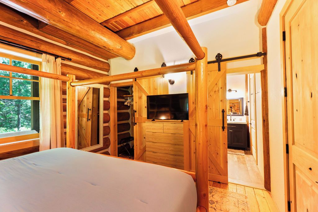 Interior de una cabaña de madera con una cama, televisor, puerta corredera y una pequeña cocina.