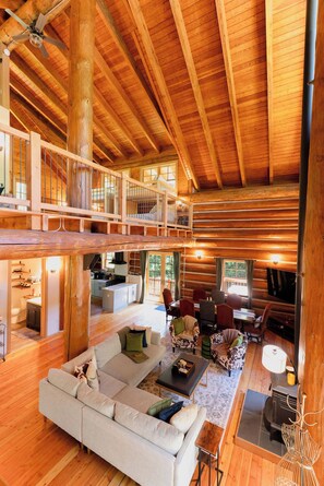Un intérieur spacieux de chalet en bois, avec un plafond haut, un coin repas, un salon doté d’un canapé et de fauteuils, et un espace cuisine.