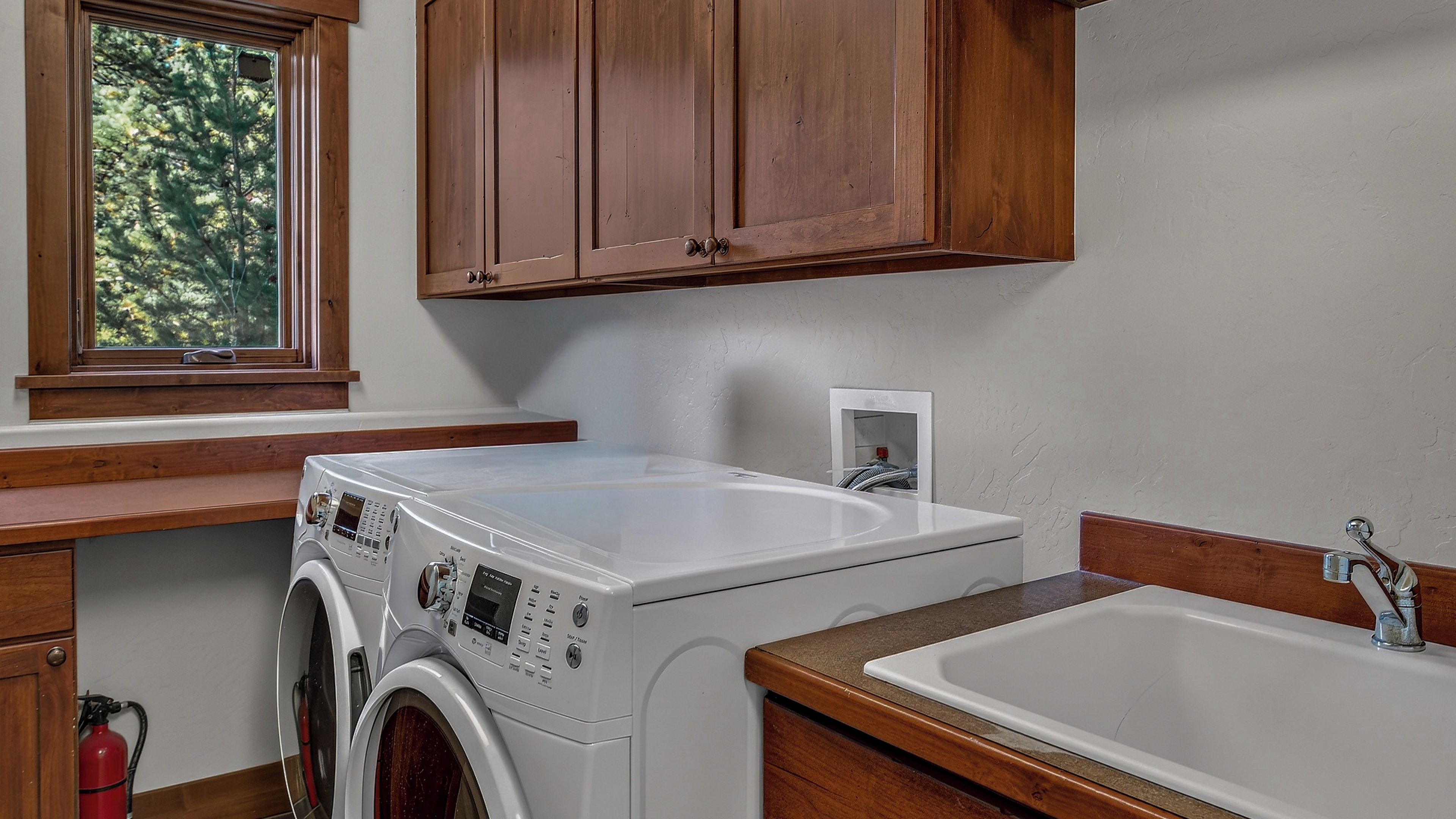 A laundry area with a washing machine, a sink, wooden cabinets, and a window with a view of trees.
