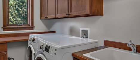 A laundry area with a washing machine, a sink, wooden cabinets, and a window with a view of trees.