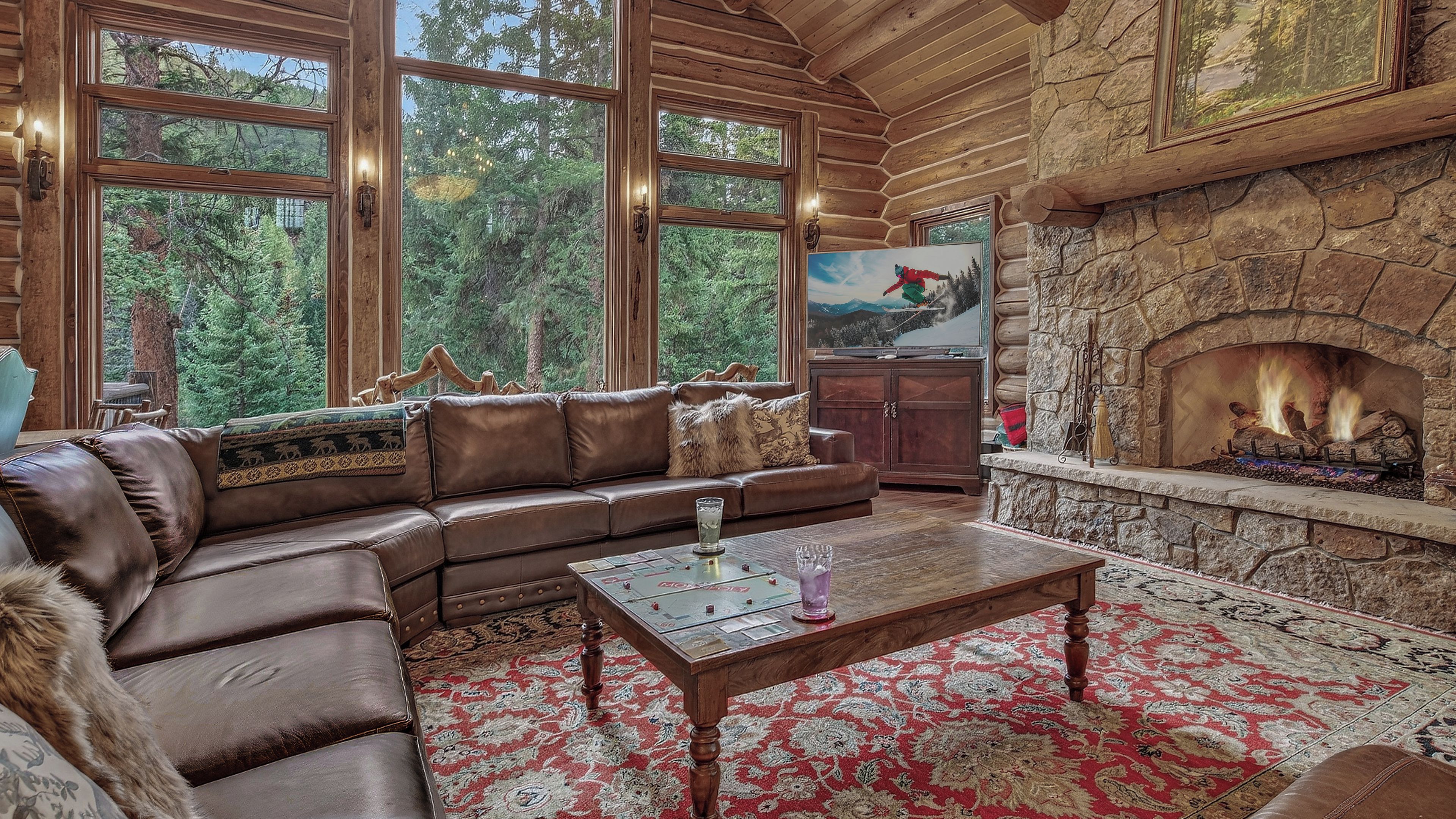 A log cabin interior with a stone fireplace, leather sofa, and large windows overlooking a forest.