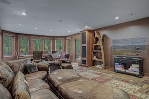 A spacious living room with leather furniture, a large window overlooking a forest, and a mounted television. - Luxury w/ River Views + Hot Tub + Private Shuttle - Rivertree Lodge (Breckenridge)