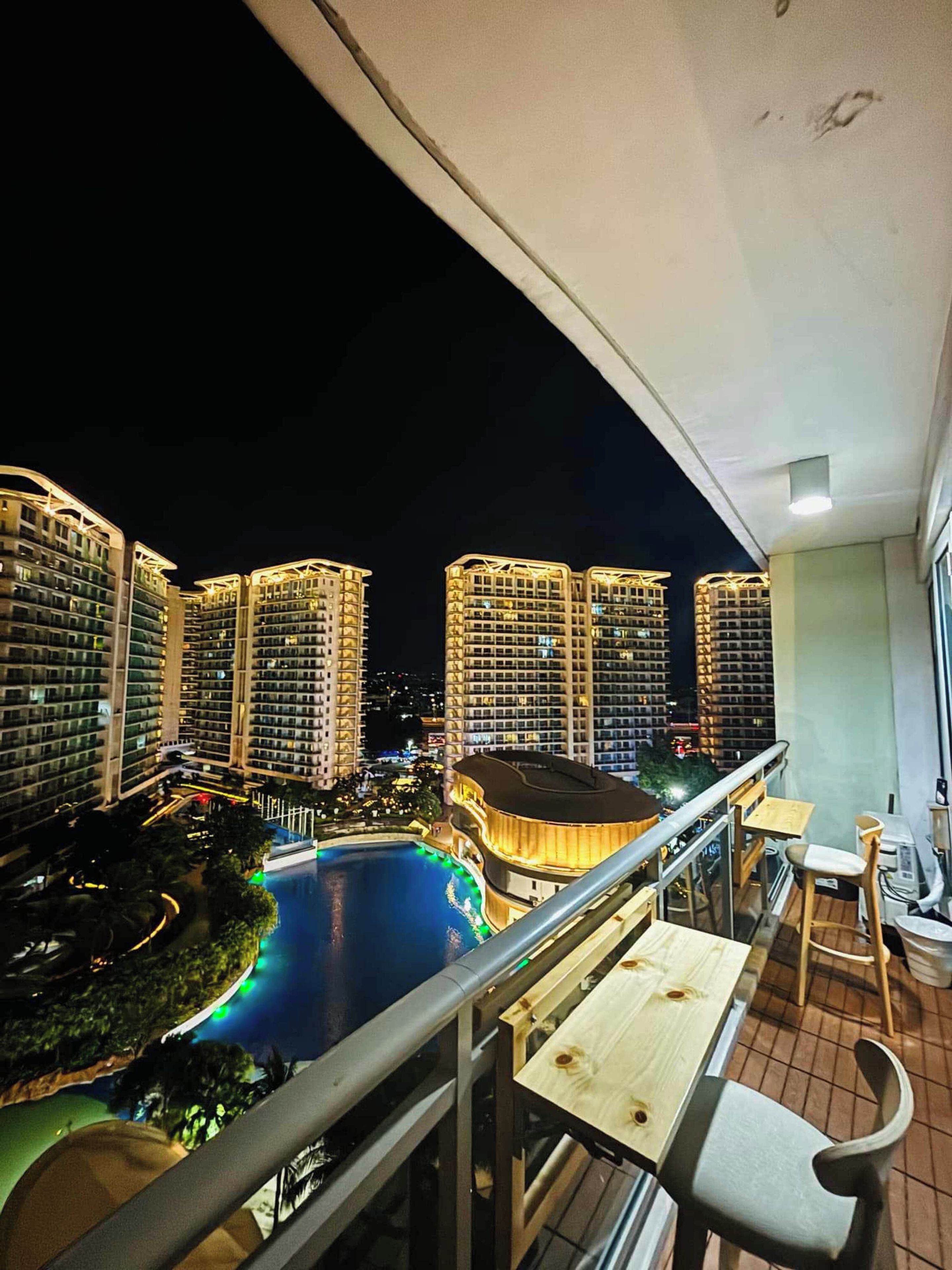 A balcony with a view of high-rise buildings and a swimming pool at night.