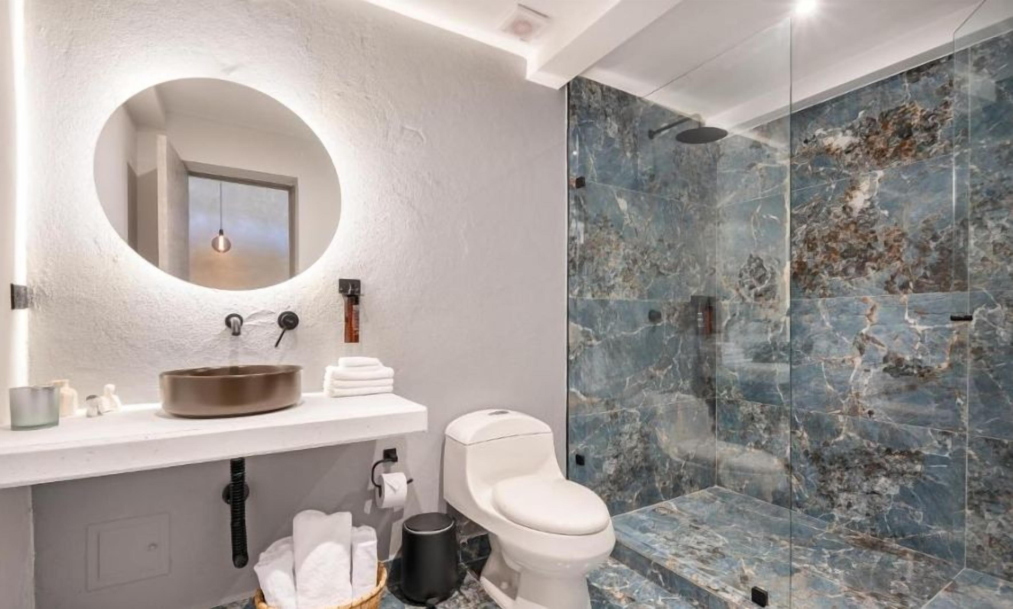 Un baño moderno con un espejo redondo, un lavabo blanco y una ducha con cerramiento de vidrio.