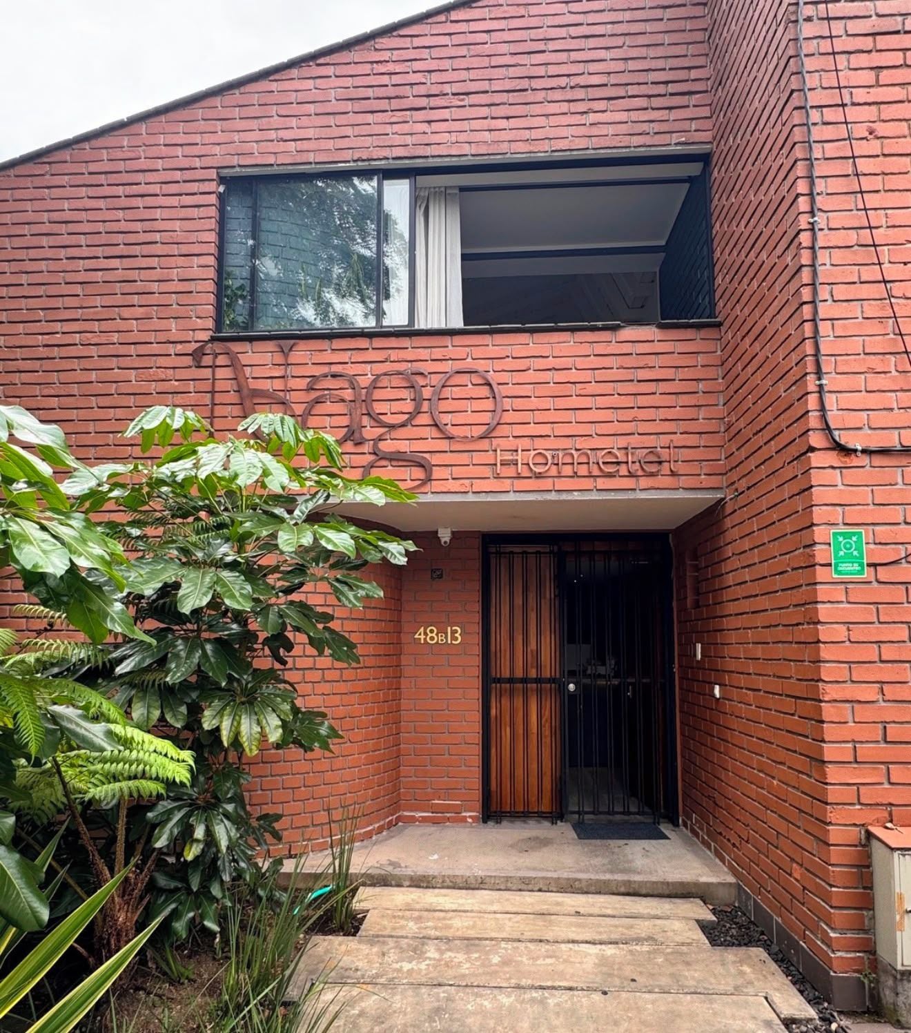 Un edificio de ladrillo rojo con un ventanal grande y una puerta. El edificio tiene un letrero que dice 'Vago Home'. Hay un letrero verde en la pared y el número '48B13' sobre la puerta.