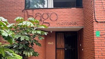 Un edificio de ladrillo rojo con un ventanal grande y una puerta. El edificio tiene un letrero que dice 'Vago Home'. Hay un letrero verde en la pared y el número '48B13' sobre la puerta.