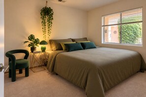 A bedroom with a bed, a chair, a bedside table with a lamp, and a hanging plant.