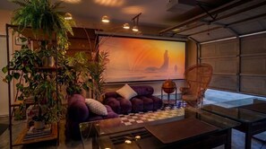 A modern living room with a purple sofa, a glass coffee table, and a large painting of a sunset.