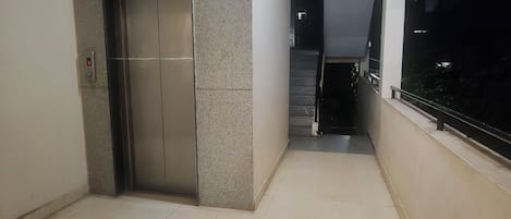 Ein Flur mit gefliestem Boden, einem geschlossenen Aufzug und einer Treppe, die zu einer weiteren Ebene führt.