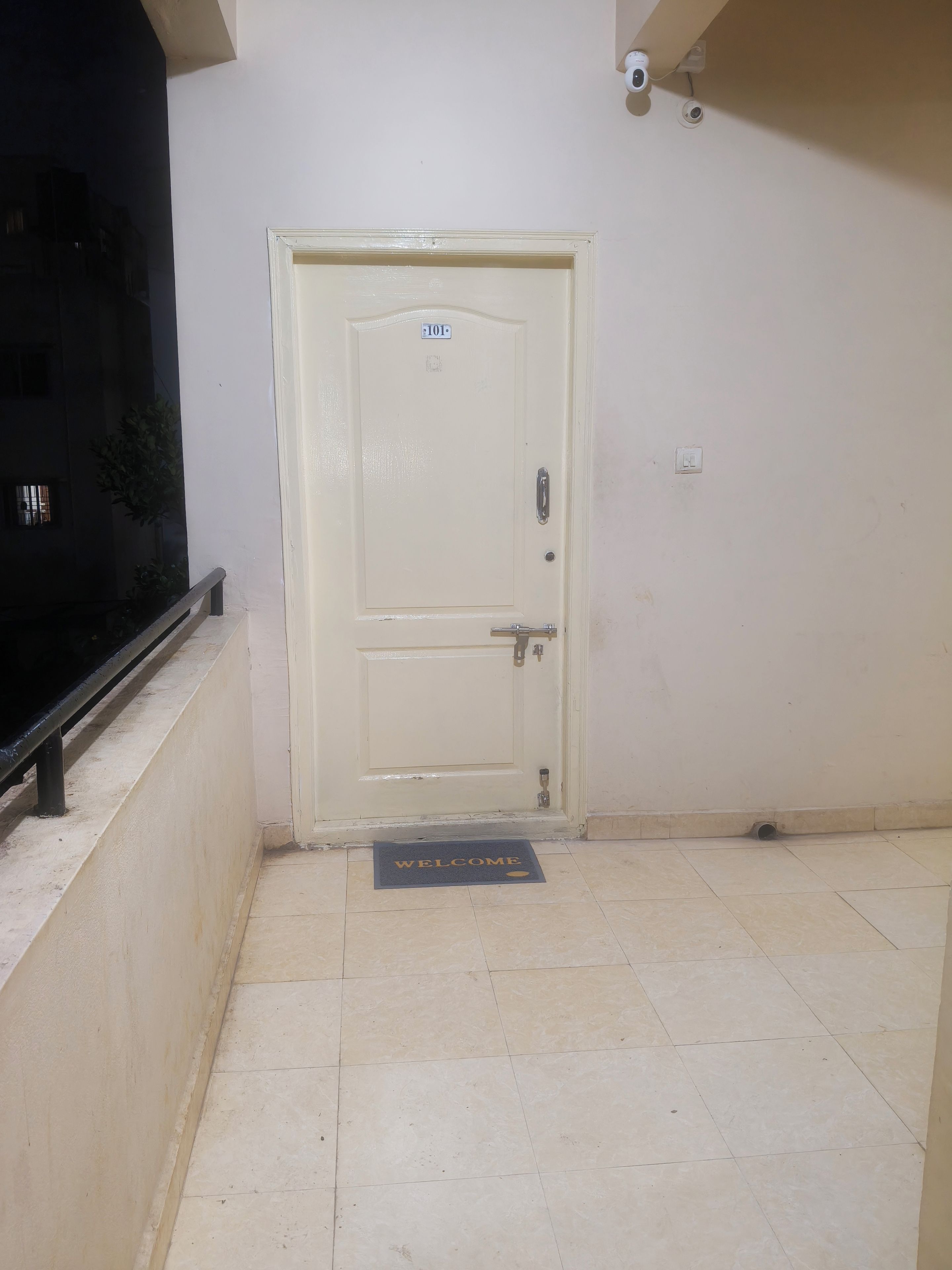 A white door with a number plate, a doormat with 'WELCOME' text, and a security camera on the wall.