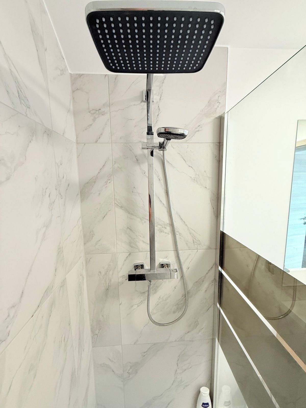 Une douche équipée d’un pommeau de douche à main et d’un panneau de commande fixé au mur, le tout installé sur un mur recouvert de carreaux de marbre.