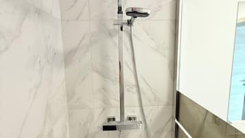 Une douche équipée d’un pommeau de douche à main et d’un panneau de commande fixé au mur, le tout installé sur un mur recouvert de carreaux de marbre.