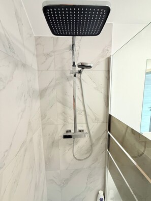 A shower with a handheld showerhead and a wall-mounted control panel, set against a marble-tiled wall.