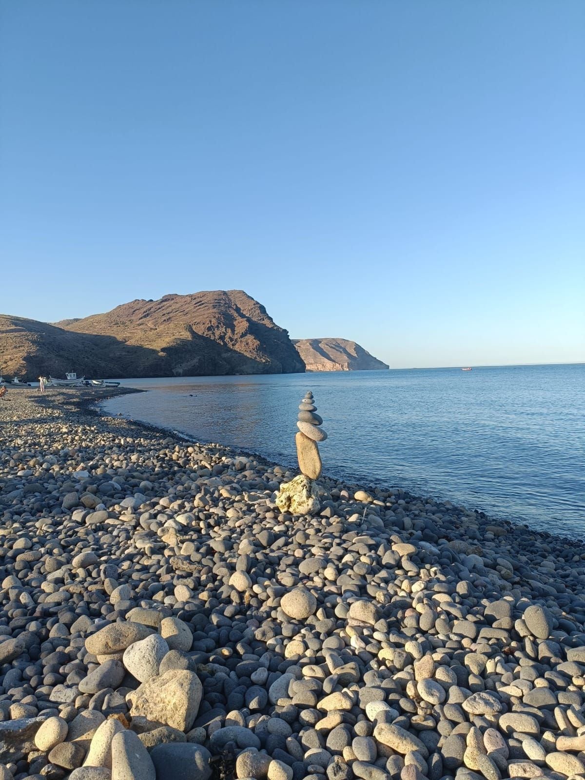 A pebble beach with a stack of stones in the foreground and boats in the background.