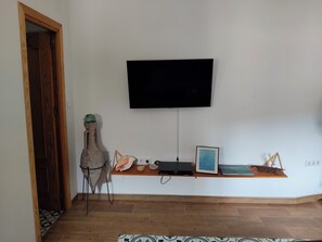 Quarto com televisão de tela plana na parede, prateleira de madeira com objetos decorativos, tapete estampado e porta de madeira.