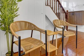 Deux fauteuils en osier avec des armatures en métal noir et une petite table en bois, placés dans une pièce dont le sol est recouvert de planches de bois, avec un escalier en arrière-plan.