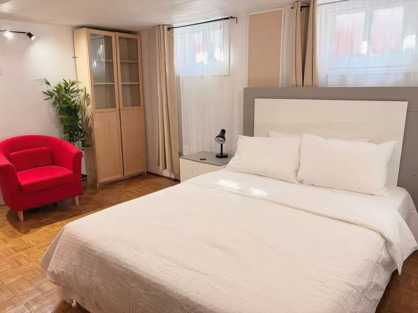 A bedroom with a bed, a red chair, a wardrobe, and a nightstand with a lamp.