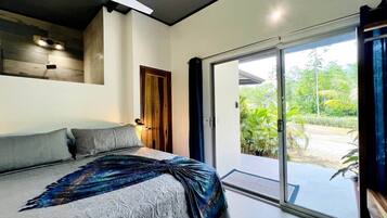Een slaapkamer met een bed, een plafondventilator, een houten deur en een schuifdeur van glas die uitkomt op een tuin.
