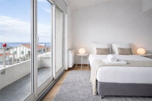A modern bedroom with a large window, a bed with pillows, a bedside table with a lamp, and a view of the sea. - [ins] igneous (Madalena)