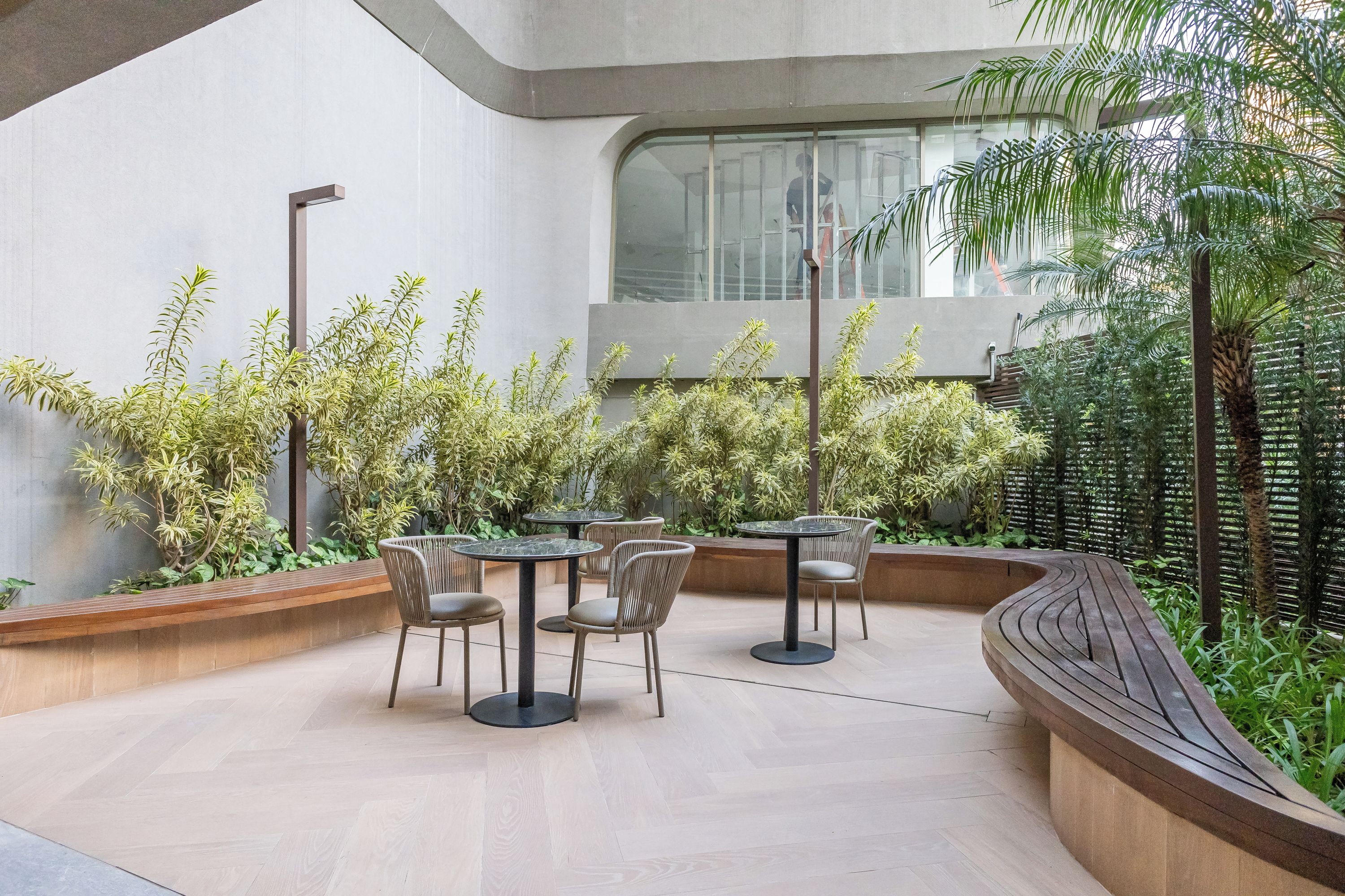 An outdoor seating area with round tables and chairs, surrounded by lush greenery and a curved wooden bench.