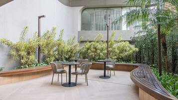 Un área de estar al aire libre con mesas y sillas redondas, rodeada de abundante vegetación y un banco de madera curvo.