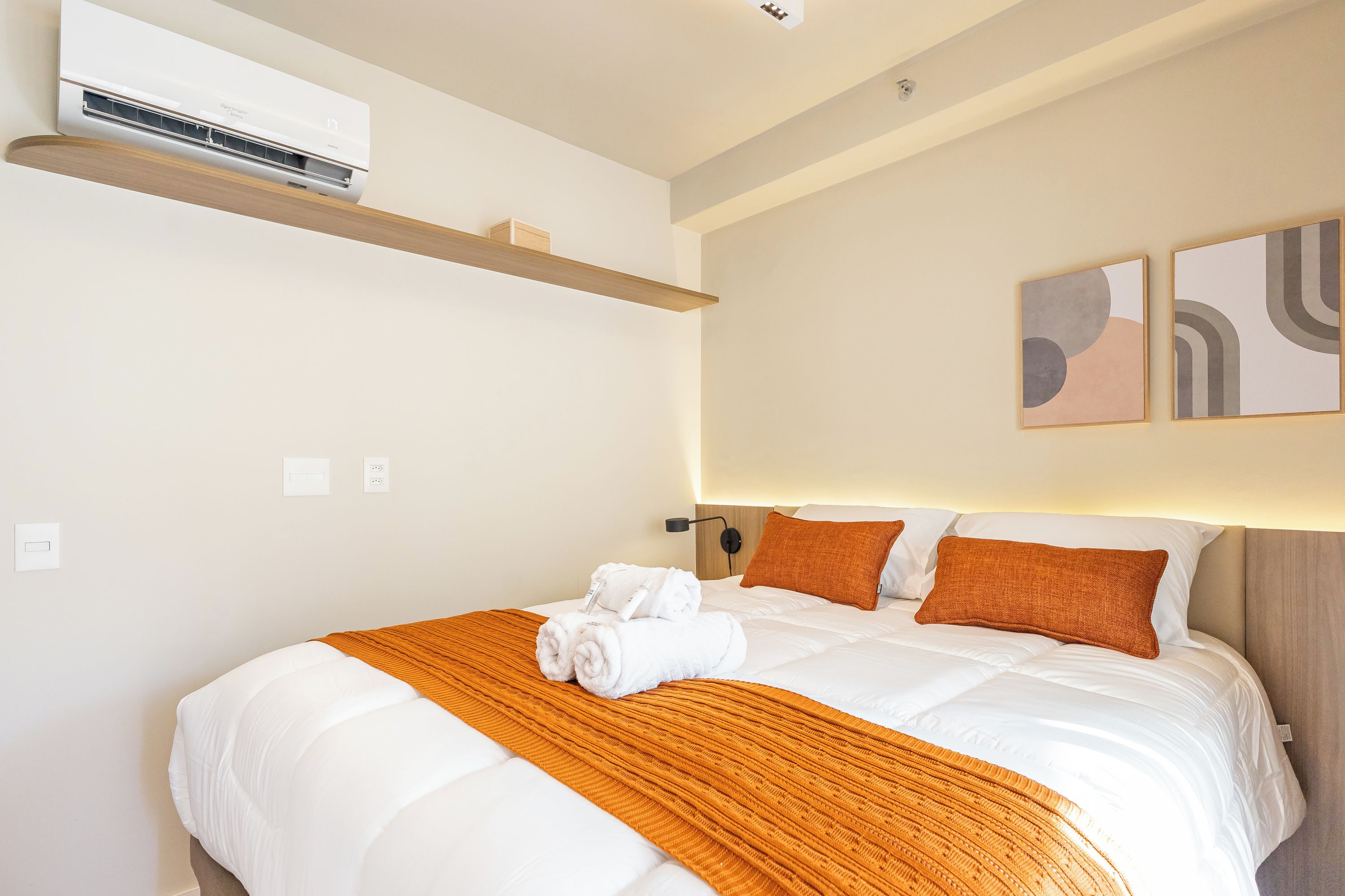 A hotel room with a bed, a wall-mounted air conditioner, a shelf, and abstract wall art.
