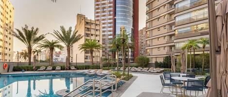 Un área de piscina con sillones de descanso y sombrillas, rodeada de palmeras y edificios modernos.