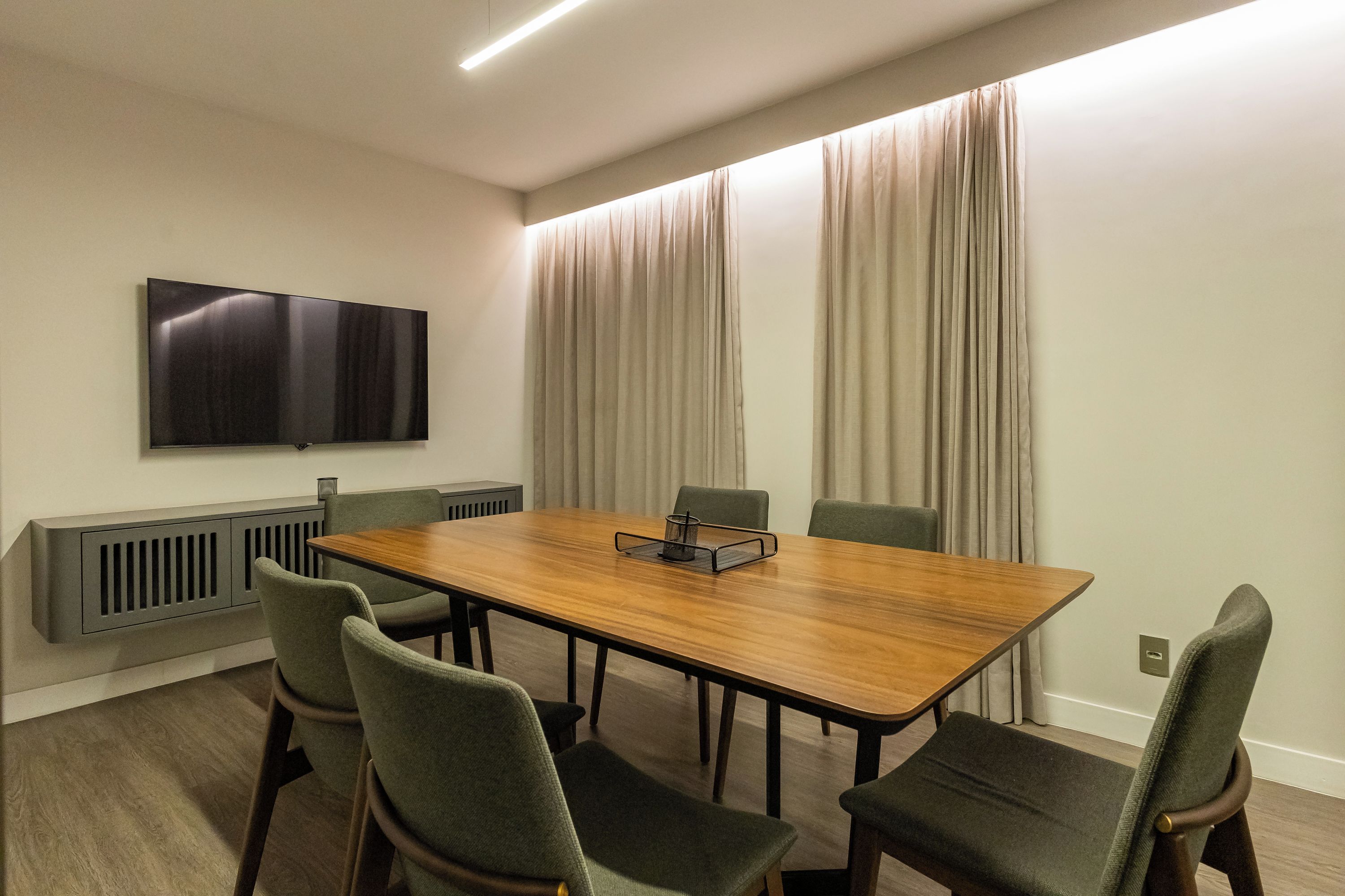 A conference room with a long table, four chairs, a flat-screen TV, and a window with curtains.