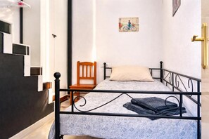 Ein Einzelbett mit gemustertem Überwurf, ein Holzstuhl, ein kleiner Tisch und ein an der Wand hängendes Bild.