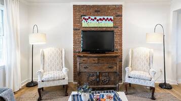 A living room with a brick fireplace, two patterned armchairs, a coffee table with a magazine, and a flat-screen TV above the fireplace.