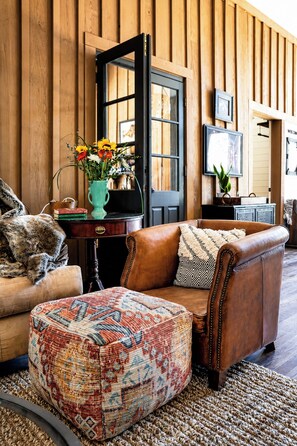 Un salón acogedor con un sofá de cuero, un reposapiés estampado, una puerta de madera y un jarrón con flores.