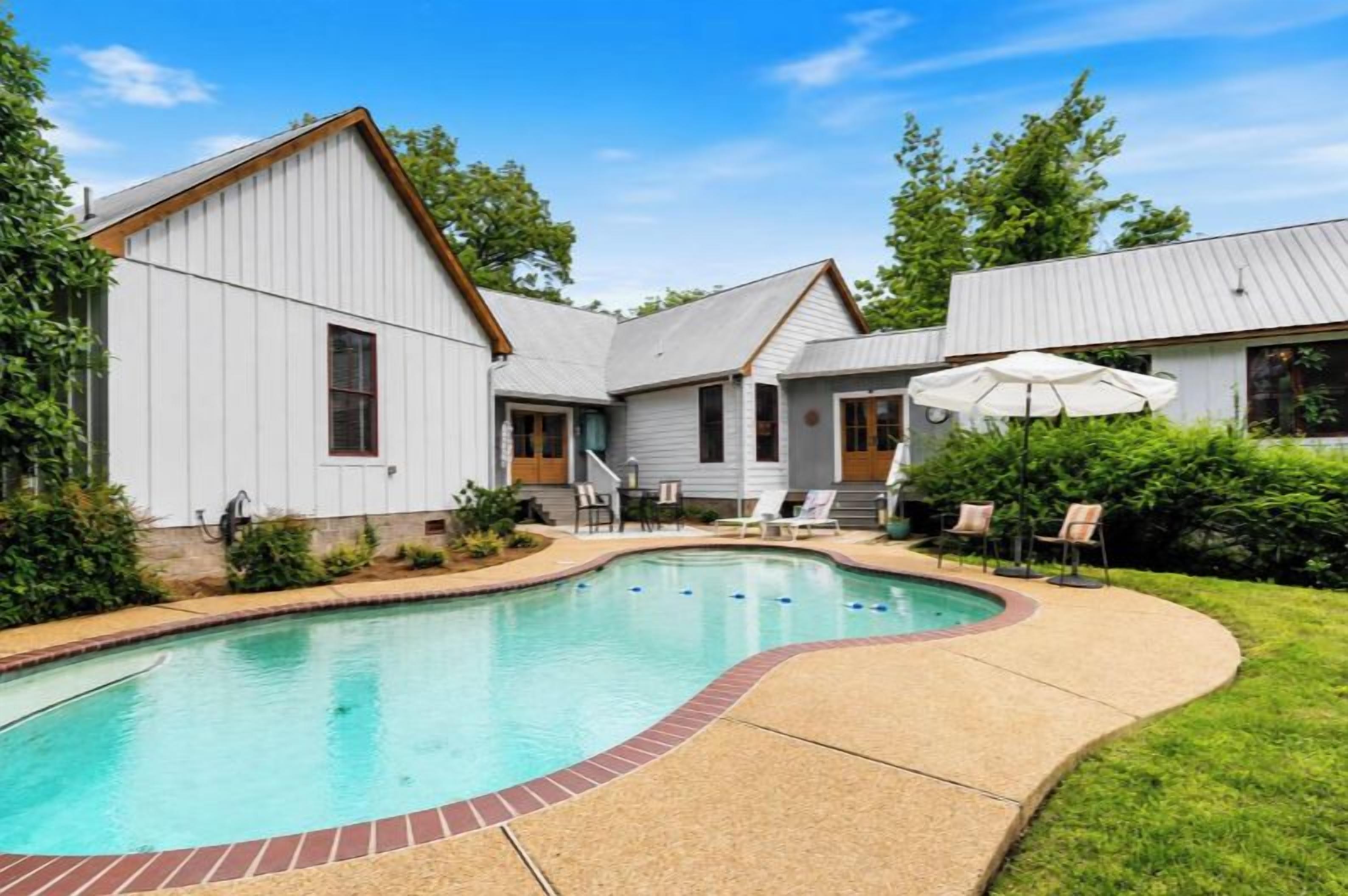 A swimming pool with a brick border, surrounded by a grassy area and a building with a sloped roof.