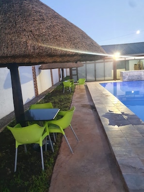Zona junto a la piscina con techo de paja, sillas verdes y piscina con azulejos.