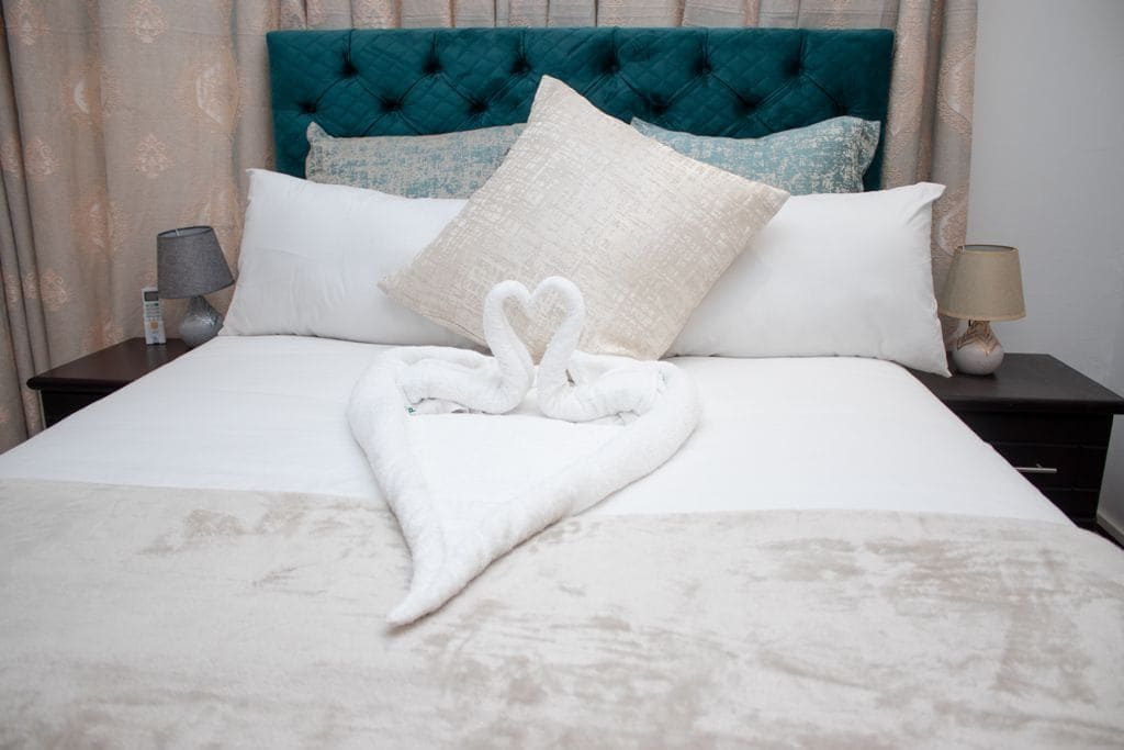 Un lit bien fait, agrémenté d’une serviette pliée en forme de cœur, d’une tête de lit turquoise et d’une lampe sur la table de chevet.