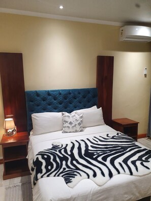 Een hotelkamer met een gebobbeld hoofdeinde, een deken met luipaardprint en een wit kussen met luipaardprint, plus een bedlampje.