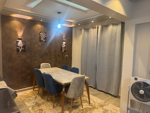 A dining area with a table and chairs, a wall-mounted light fixture, and a ceiling fan. - Modern 2BR in 6th October Balcony, Parking, WiFi & Smart TV (6th October)